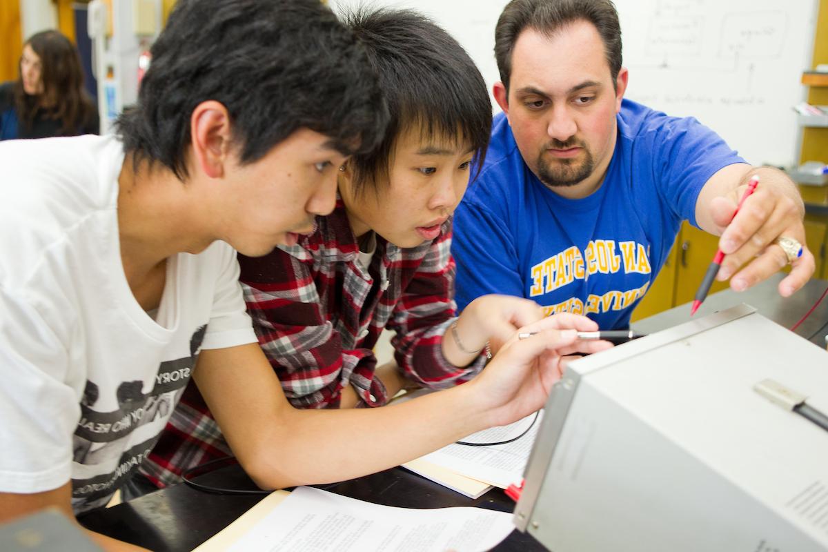 Three students gather around a monitor and point to it with their pencils.